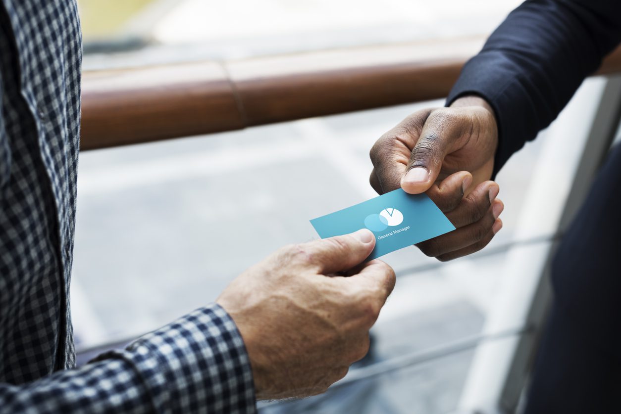 Are business cards still relevant? For millions of people, the answer is yes. An image of two hands exchanging a dark turquoise business card with a minimalist logo.